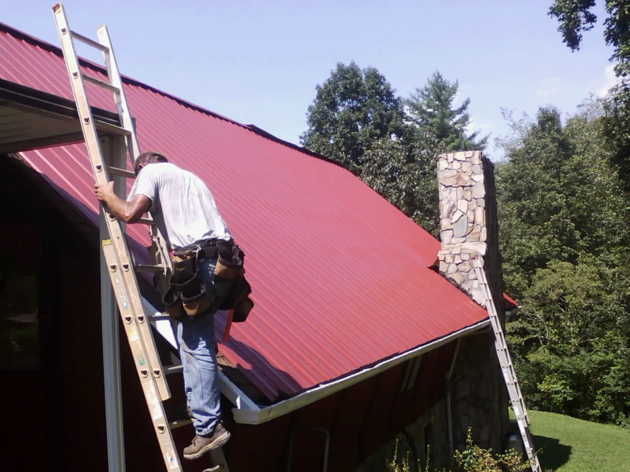 Roofer on a ladder beside a red metal roof and stone chimney — Soffit & Fascia Repair by Roofing Brothers in Grasston