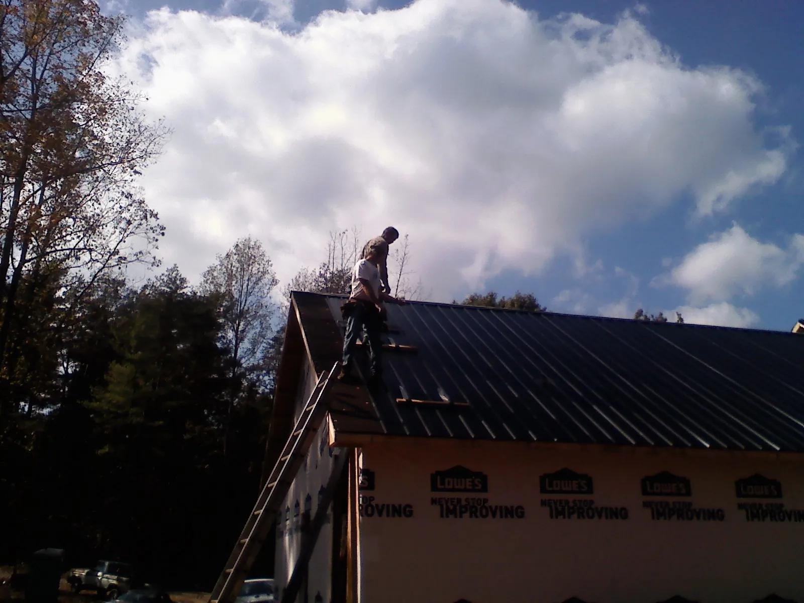 Roofer fastening dark metal panels high against a cloudy sky, from a Roof Inspection job in Grasston